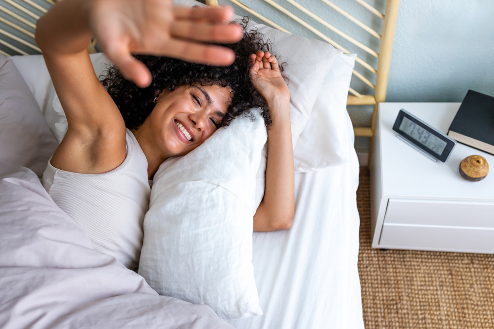 Young woman smiling while waking up in bed, shielding her eyes from morning sunlight