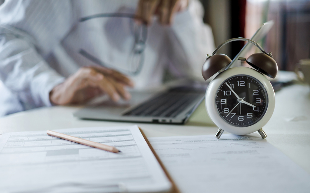 Selective focus on alarm clock, Asian businessman working with laptop and document on table before work time end in a day, time management concept, routine work, office hour